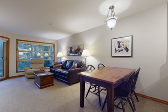 The living area within Aspens on Blackcomb Unit 415 features a comfortable dark leather sofa and a recliner, alongside a wooden coffee table and a dining table with chairs, all set on a light-colored carpet.