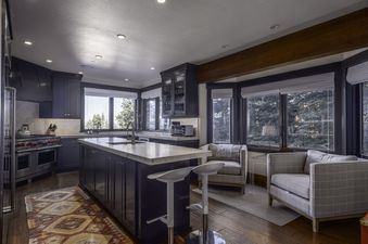 A modern kitchen with dark cabinetry, a large island, and a sitting area with plaid armchairs, all featuring expansive windows with views of evergreen trees.