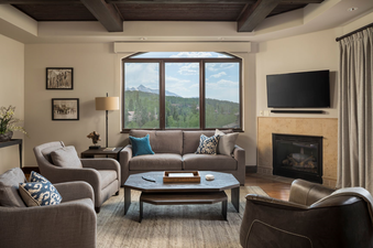 The living room of Lumiere Three Bedroom Luxury features a comfy couch and armchairs, a large window overlooking stunning mountain views, and a fireplace.