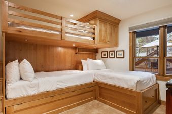 A guest bedroom in Montaneros 212 featuring a custom-built light wood bunk bed system with three twin beds, white bedding, built-in storage, and a large window.