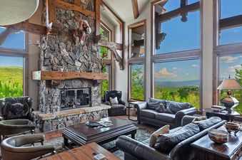 A spacious living room featuring a grand stone fireplace, comfortable seating, and large windows offering panoramic mountain views.