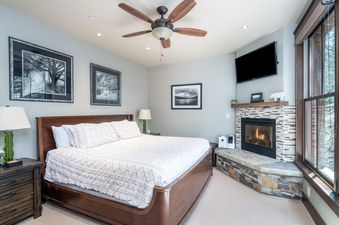 A cozy bedroom in Trails Edge 8 featuring a king bed, corner stone fireplace with a fire lit, and natural light from large windows.