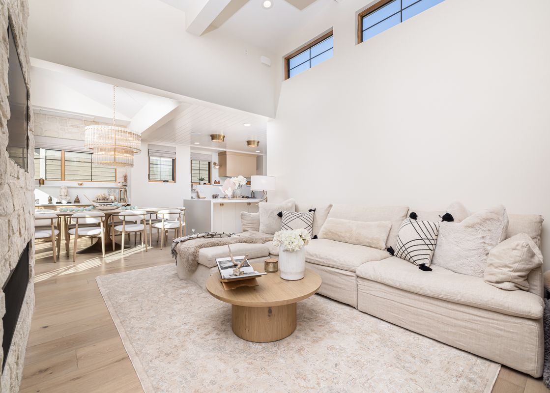 A bright and open-concept living room with a large L-shaped sofa, a round coffee table, and a stone fireplace, leading to a dining area and kitchen.