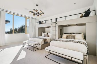 A modern bunk room in Founders Place 1704 with two built-in bunk beds, plush white bedding, a contemporary chandelier, and large windows offering views of pine trees.