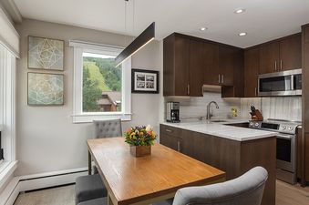 A modern dining and kitchen area with a wooden table, gray chairs, dark wood cabinetry, and stainless steel appliances.