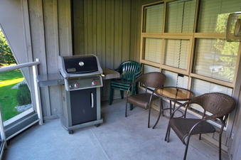 The image shows a balcony with a barbecue grill and outdoor furniture, offering a view of the grassy area and trees beyond.