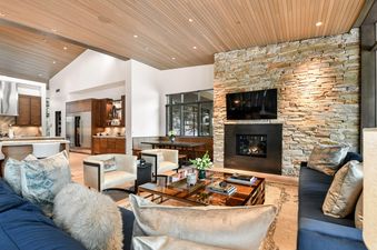 A spacious, open-plan living room with vaulted wood-paneled ceilings, a stone fireplace, a large TV, comfortable seating, and a view of the kitchen and bar area.