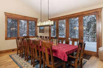 The dining room at Last Nickel Lodge features a long wooden table with a red tablecloth, surrounded by wooden chairs, and large windows offering a snowy outdoor view.