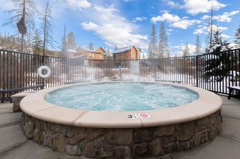 An inviting outdoor hot tub at The Aerie steams gently under a bright blue winter sky, surrounded by snow-dusted trees and rustic buildings.