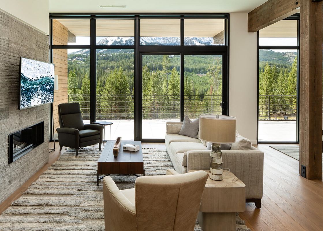 The interior of Moonlight Basin 212 Lakelodge, featuring a large window with a view of the forested mountains, a fireplace with a black hearth, a white sofa and a large, brown leather armchair, a wooden coffee table and side table, and a beige rug.