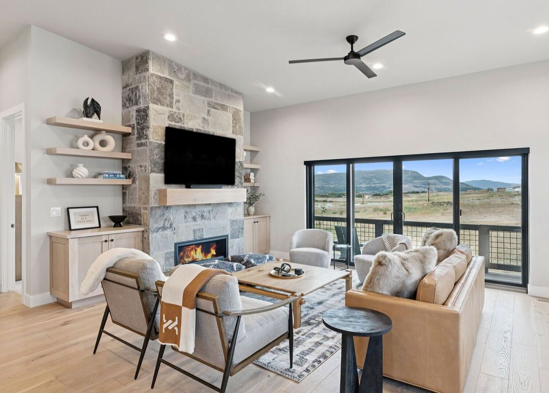 A spacious living room with a stone fireplace, light wood furniture, a tan leather sofa, and large sliding glass doors revealing a mountain view.