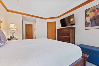 A cozy bedroom with a large bed, nightstand with a lamp, a dresser with a TV mounted above it, and two wooden doors.