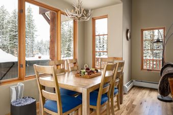 The dining room at Moonlight Mountain 38B Cowboy Heaven features a large wooden table with blue-cushioned chairs, an antler chandelier, and expansive windows showcasing a snowy forest and an outdoor hot tub.
