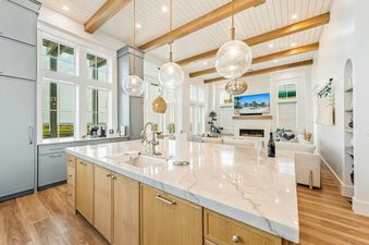 A modern kitchen with a large marble island and light wood cabinets, open to a living room with a fireplace and ocean views.
