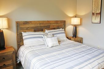 The Meadow Ridge 8 Unit 6 features a bedroom with a cozy bed, adorned with light blue and white striped bedding, and flanked by two bedside tables each with a lit lamp.