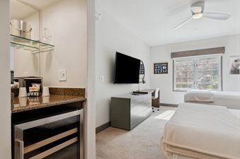 A bedroom in The Charter F235 featuring two twin beds, a wall-mounted television, a desk with a chair and lamp, and a wet bar with a mini-fridge and coffee machine.
