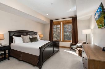 The primary bedroom in Town Pointe C304 features a dark wood bed with white linens, two windows with wooden frames and dark brown curtains offering views of the village, and a large flat-screen TV.