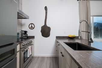 A kitchen with stainless steel appliances, a large wooden cutting board hanging on the wall, and a view of the mountains outside the window.