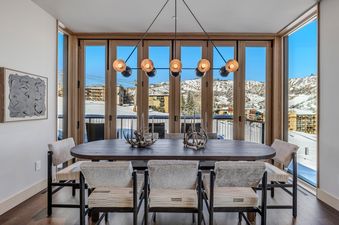 A modern dining room with a long wooden table, faux fur chairs, and a contemporary chandelier, set against a backdrop of snow-covered mountains seen through large windows.