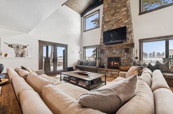 The Mission View Escape's living area showcases a floor-to-ceiling stone fireplace with a roaring fire, two large, comfortable sofas, and a wooden coffee table, all set against a backdrop of vaulted ceilings and expansive windows that offer stunning views