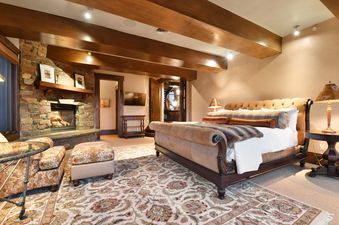 A luxurious bedroom at Ski Dream Home features a grand upholstered bed, a stone fireplace, and elegant wooden ceiling beams, creating a warm and inviting atmosphere.