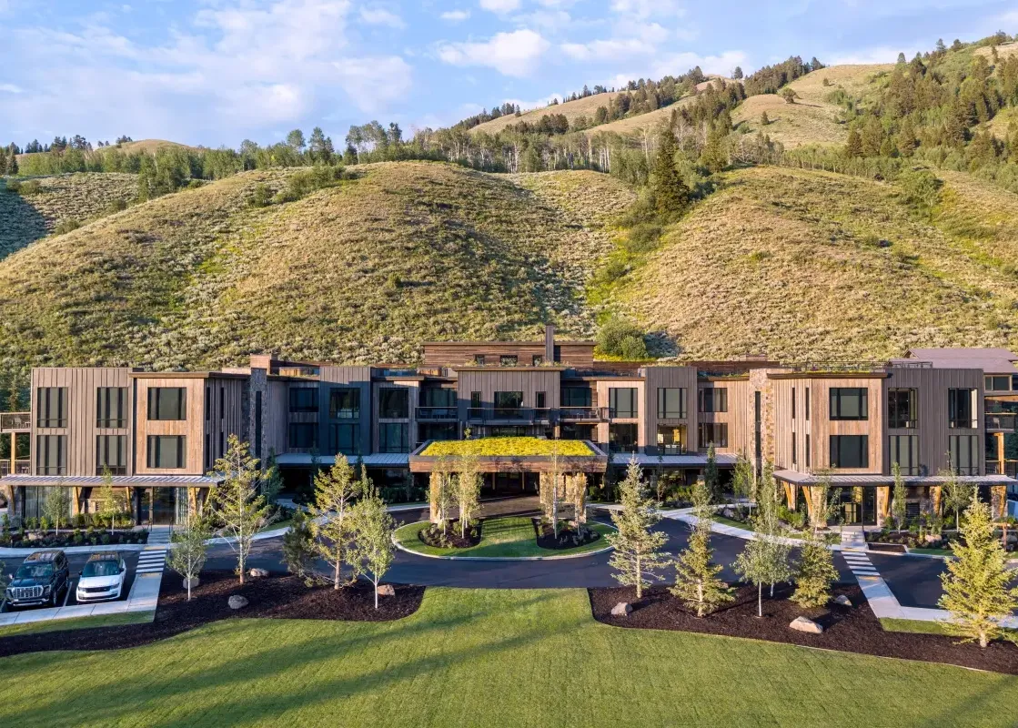 A modern, multi-story lodge with a green roof and a circular driveway, set against a backdrop of lush green hills and a blue sky.
