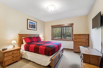 The bedroom of Pines Unit 120 features a bed with a red and black plaid comforter, a wooden headboard, two nightstands with lamps, a window with an exterior view of trees, a tall wooden dresser, and a wall-mounted TV.