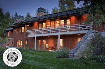 A rustic-modern wooden home named Overlook Clear Creek is illuminated at twilight, featuring a multi-level structure with glowing windows, a long balcony, stone chimneys, and a pet-friendly seal on the lawn.