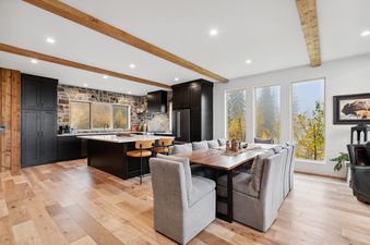 An expansive open-concept room featuring a modern kitchen with black cabinets and a dining area with a large wooden table and upholstered chairs, all illuminated by natural light and recessed ceiling lights.
