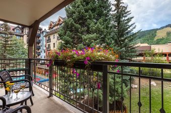 A vibrant balcony at Montaneros 214 adorned with colorful flowers, featuring outdoor seating and offering views of surrounding buildings, lush pine trees, and a distant mountain slope under a cloudy sky.