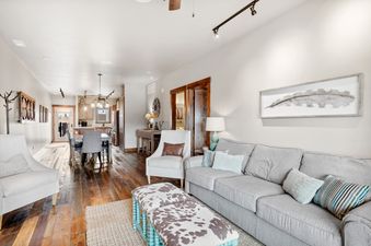 A spacious, open-concept living room with wooden floors, a gray sofa, a cowhide ottoman, and an adjacent dining area and kitchen.
