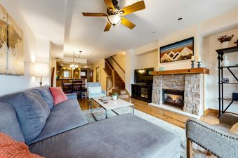 A living room with a gray sectional couch, a coffee table, a fireplace, a television, a staircase, and a dining room in the background.