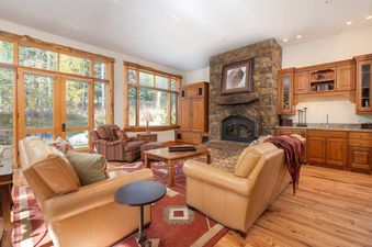 A cozy, rustic living room at Granite Ridge Lodge 6 with comfortable seating, a stone fireplace, and views of the surrounding woods.