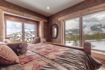 A rustic bedroom with a large bed, patterned blankets, and pillows, offering expansive window views of snow-capped mountains and pine forests under a cloudy sky.