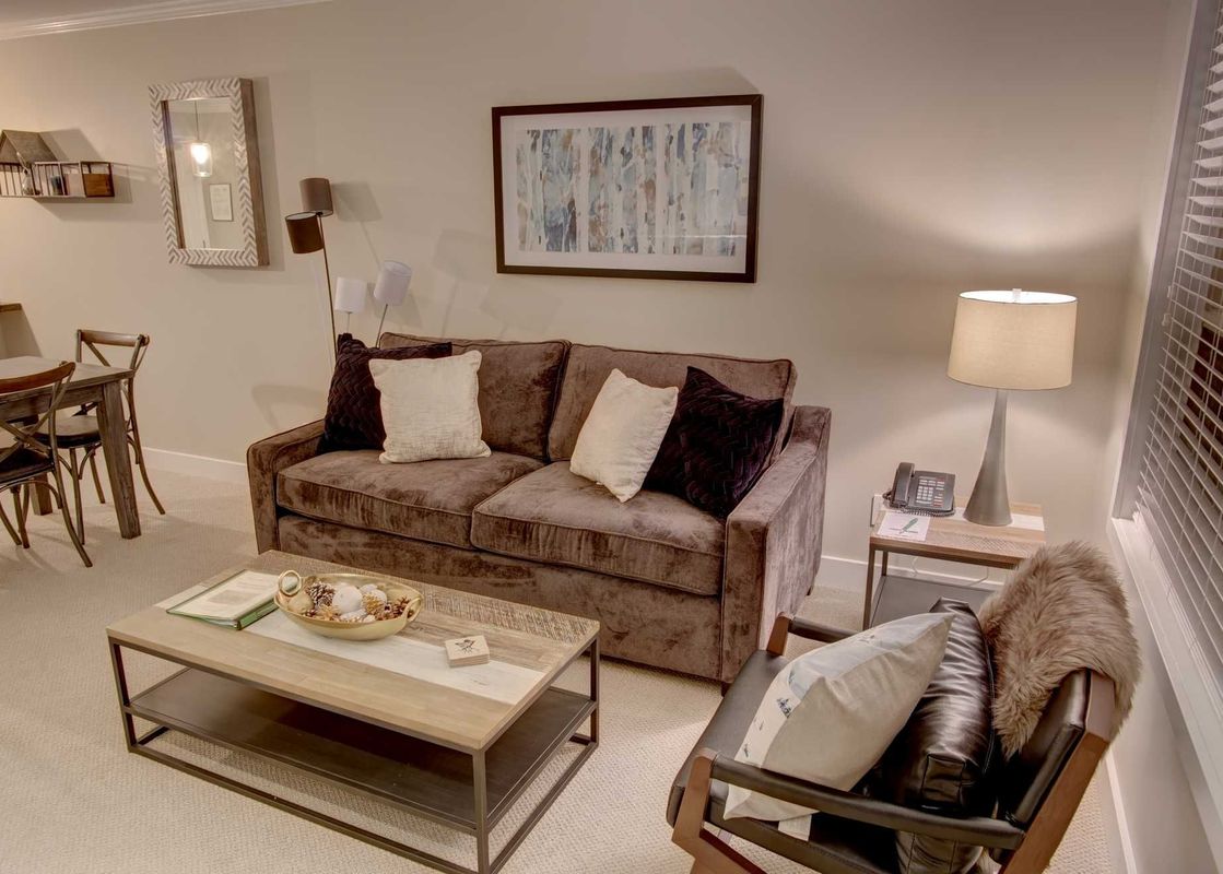 The inviting lounge area in Aspens on Blackcomb Unit 304 features a plush brown sofa adorned with white and dark pillows, a stylish coffee table, and an armchair, all resting on soft, light-colored carpeting.