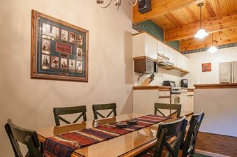 The dining area at Kutuk 201 features a polished wooden table with six chairs and a colorful table runner, illuminated by overhead pendant lights, with the kitchen visible in the background.