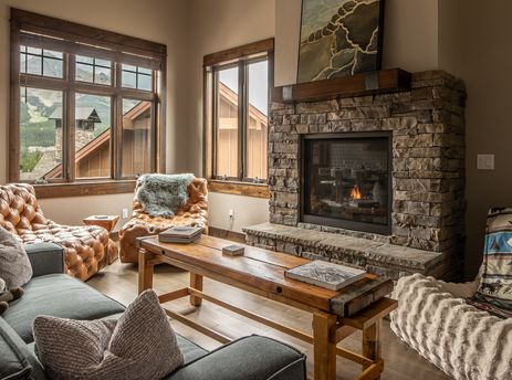 A cozy living room in Alpenglow Condo 26D with a stone fireplace, plush seating, and large windows overlooking snowy mountains.