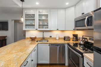 The kitchen in Villa Valhalla 2 Bedroom Platinum Unit 2 features a spacious granite countertop, stainless steel appliances including a microwave, dishwasher, and refrigerator, and white cabinetry with glass inserts.