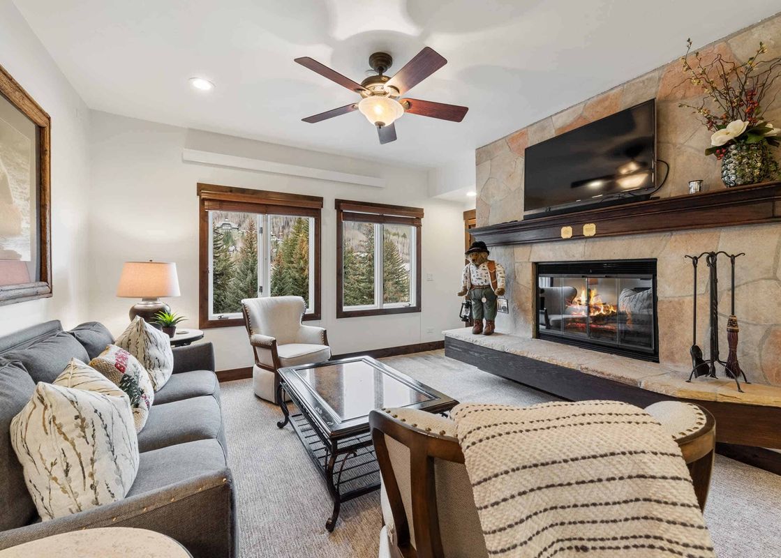 A cozy living room in The Charter D3205 with a stone fireplace, a gray sofa with decorative pillows, a glass coffee table, and large windows looking out onto snowy pine trees.