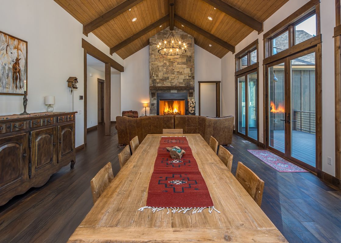 The dining area and living room of the Stony Creek Chalet feature a long wooden dining table, a stone fireplace, and large glass doors leading to a deck.