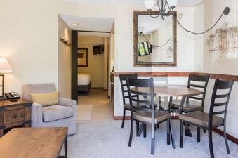 A cozy dining area with a round table and four chairs is visible, leading into a living space with an armchair and a glimpse of a bedroom beyond.