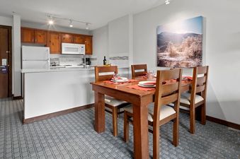 The dining area and kitchen of the Blackcomb Springs Two Bedroom Slopeside Suite are visible, featuring a wooden dining table set for four, adjacent to a kitchen with white appliances and light wood cabinetry.