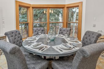 A round dining table with six upholstered chairs and full place settings, surrounded by bay windows offering a view of trees.