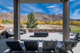 A luxurious outdoor patio at Big Wood River Retreat features comfortable lounge seating and a sleek fire pit, framed by wooden pillars with a stunning view of autumn-colored trees and mountains under a blue sky.