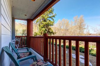 A serene private balcony at Snow Flower 012 with green Adirondack chairs and rustic wooden railing, offering tranquil mountain views with patches of snow and lush greenery under a clear blue sky.