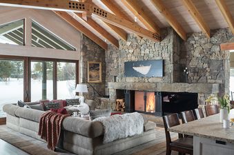 A cozy living room at The Paintbrush Retreat features a stone fireplace, exposed wooden beams, a large sectional sofa, and expansive windows showcasing a snowy outdoor scene.