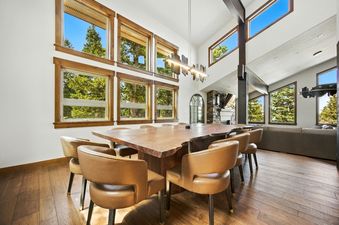 A spacious dining area featuring a large live-edge wooden table, leather chairs, and multiple windows offering views of evergreen trees.