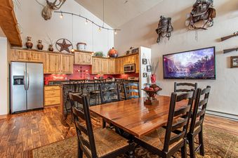 A spacious kitchen with a dining table and chairs, a large stainless steel refrigerator, and a flatscreen TV in the background.