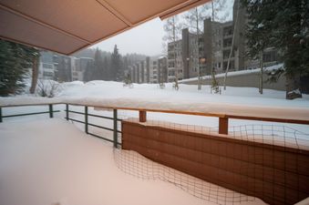 A snowy winter scene from the perspective of a balcony, with a vast snow-covered field leading to multistory buildings and pine trees under a cloudy sky.
