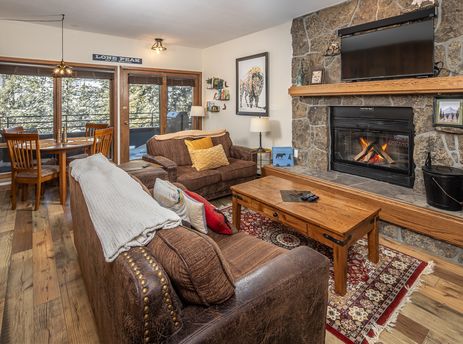 A cozy living room at the Snowflake at Skycrest features leather sofas, a wooden coffee table, a stone fireplace, a TV, and a dining area with scenic winter views.